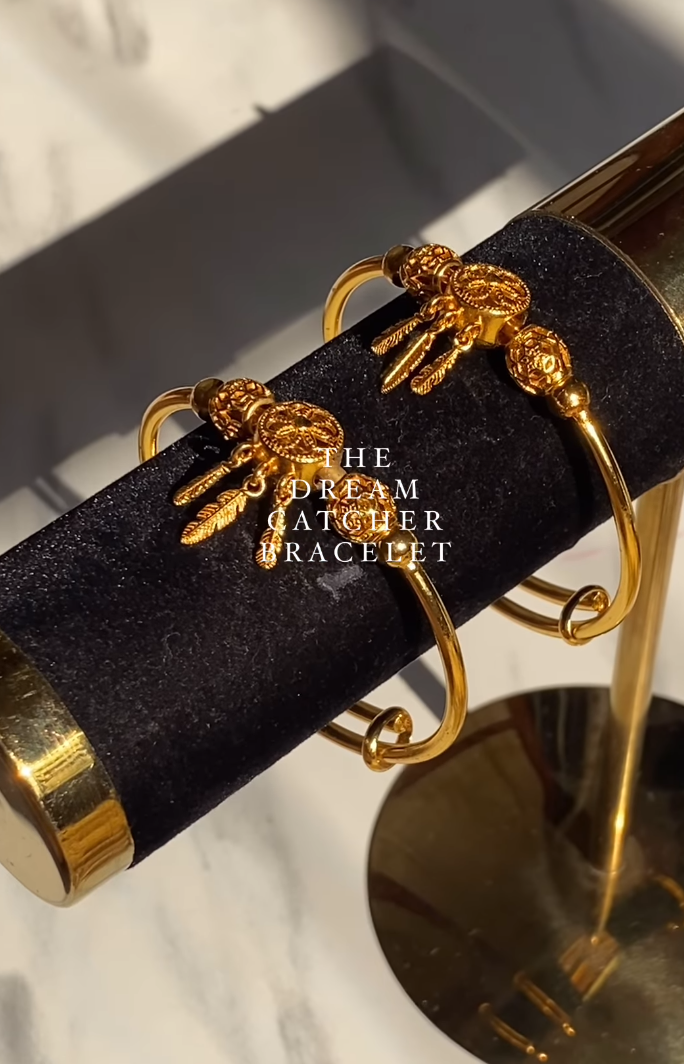 A gold-colored bracelet with two hooks, displayed on a marble surface with a reflective gold background.