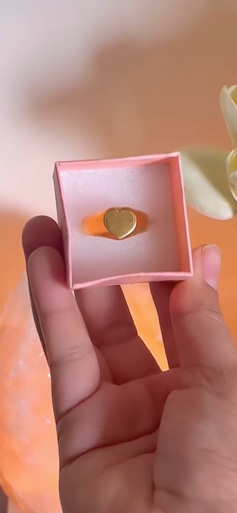 Heart-shaped gold ring in a pink box held by a hand against an orange background