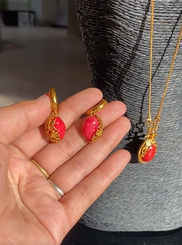 A person holding a small red and gold earring and necklace set, with the earrings resembling cherry blossoms.
