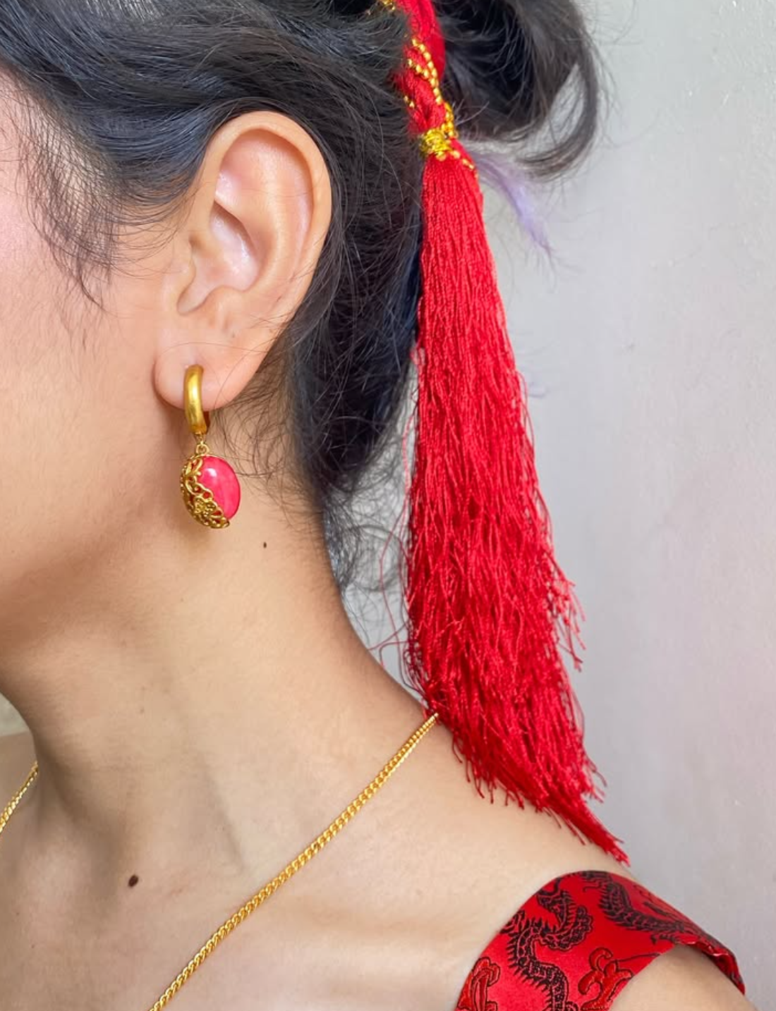 Close-up of an ear wearing a gold earring with a pink gemstone, red hair accessory, and red and black traditional outfit.