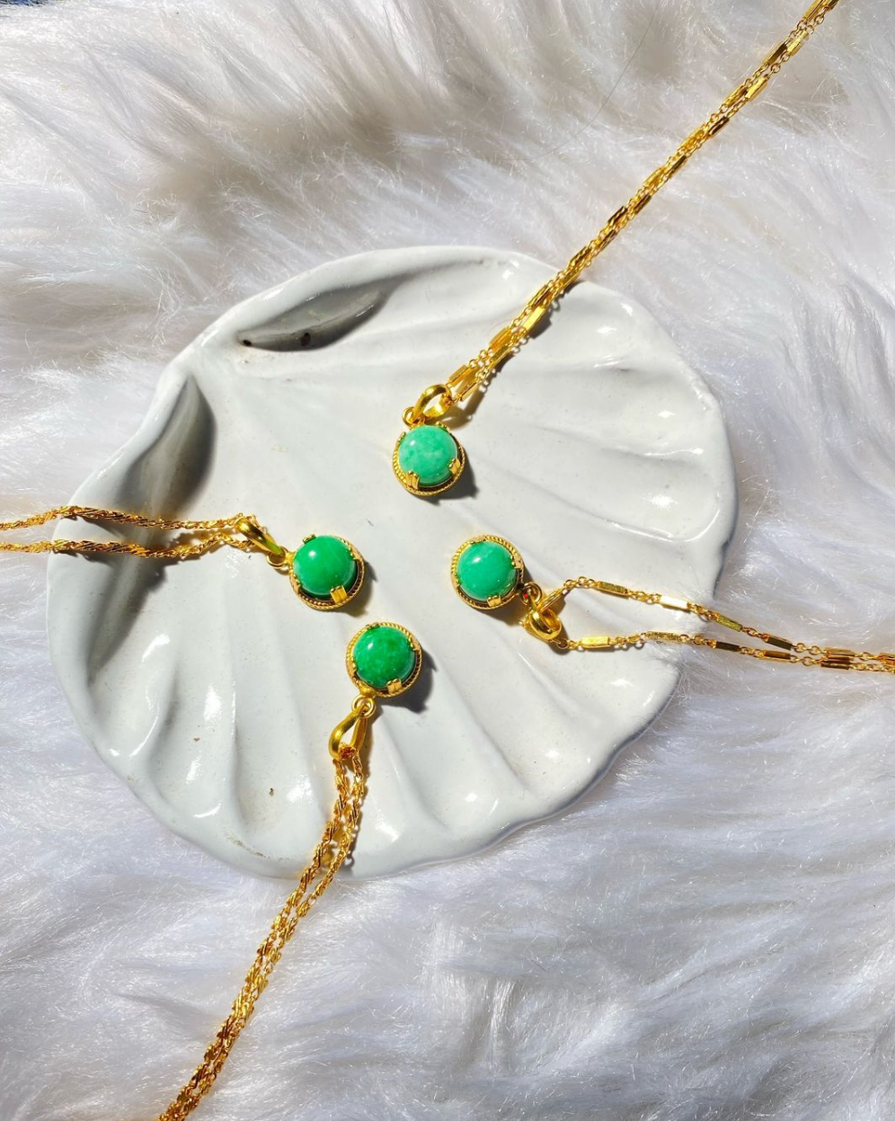 Four jade pendants displayed on a white ceramic plate, with gold-colored metal chains, set against a furry white background.