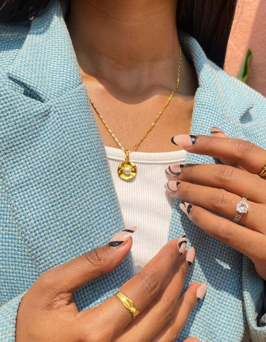 A person wearing a gold-colored oyster pendant necklace with a matching pearl earring, in a blue jacket and a white top, with their hands near their neckline.